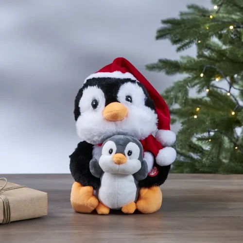 Pluszowy pingwin w czapce Mikołaja trzymający małego pingwinka, ustawiony na stole obok zielonej choinki z delikatnymi światełkami.