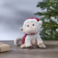 Pluszowa figurka małpki w czerwonej czapce Mikołaja i biało-czerwonym szaliku, siedząca na drewnianej powierzchni, z choinką i delikatnymi światełkami w tle.