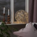 Kula LED z metalowego drutu stojąca na parapecie obok świec i roślin. Ciepłe światło widoczne w ażurowej konstrukcji buduje przytulną atmosferę wieczornego wnętrza.
