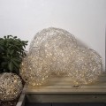 Zestaw trzech kul LED z metalowego drutu na drewnianym podwyższeniu przy roślinach. Ciepłe punkty światła widoczne w ażurowych formach budują nastrojową, dekoracyjną aranżację.