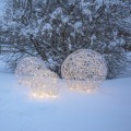 Trzy świetlne kule LED z metalowego drutu ustawione na śniegu w ogrodzie. Delikatne ciepłe światło rozświetla ażurowe formy na tle ośnieżonych krzewów, tworząc zimową, nastrojową dekorację.
