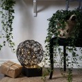 Czarna dekoracyjna kula LED z metalowego drutu ustawiona na podłodze we wnętrzu, obok zielonych roślin i zapakowanego prezentu. Ciepłe punkty światła w ażurowej formie tworzą nastrojowy, domowy akcent.