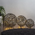 Trzy czarne kule LED z metalowego drutu ustawione na podłodze przy ścianie i roślinach. Różne rozmiary i ciepłe punkty światła w ażurowych formach budują harmonijną, przytulną kompozycję.