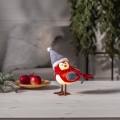 Ptaszek dekoracyjny w czerwonym stroju z szalikiem i szarą czapką, ustawiony na jasnym drewnianym stole. W tle zimowa aranżacja z gałązkami i owocami, tworząca ciepły, świąteczny klimat wnętrza.