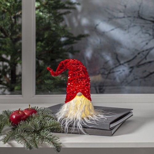 Świąteczny skrzat dekoracyjny w czerwonej, cekinowej czapce i z jasną brodą, ustawiony na parapecie obok gałązek i owoców. W tle zimowy widok za oknem, tworzący ciepłą, domową aranżację.