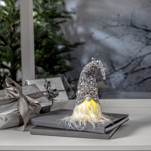 Skrzat dekoracyjny w srebrnej, cekinowej czapce i z jasną brodą, ustawiony na parapecie obok zapakowanych prezentów i gałązek. Zimowy widok za oknem oraz miękkie światło tworzą spokojną, świąteczną aranżację.