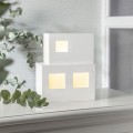 Lampa dekoracyjna dom LED w białej, geometrycznej formie z podświetlanymi oknami, ustawiona na jasnej powierzchni obok zielonej rośliny. Ciepłe światło tworzy spokojną, minimalistyczną aranżację.