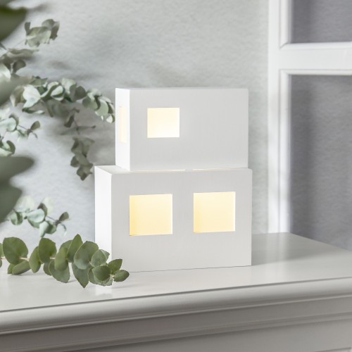 Lampa dekoracyjna dom LED w białej, geometrycznej formie z podświetlanymi oknami, ustawiona na jasnej powierzchni obok zielonej rośliny. Ciepłe światło tworzy spokojną, minimalistyczną aranżację.