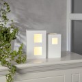 Lampa dekoracyjna dom LED w białej, geometrycznej formie z podświetlanymi oknami, ustawiona na jasnej powierzchni obok zielonej rośliny. Ciepłe światło buduje spokojną, minimalistyczną atmosferę wnętrza.
