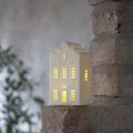 Biała ceramiczna dekoracja w kształcie domku z ciepło podświetlonymi oknami, ustawiona na ceglanej krawędzi przy ścianie; nastrojowe światło i delikatne tło.