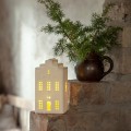 Biała ceramiczna dekoracja w kształcie domku z ciepłym światłem w oknach, ustawiona na ceglanej półce obok doniczki z zieloną gałązką; subtelne, przytulne oświetlenie wnętrza.