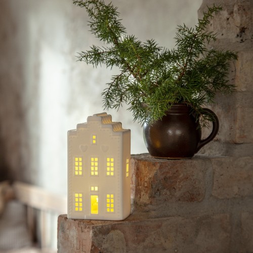 Biała ceramiczna dekoracja w kształcie domku z ciepłym światłem w oknach, ustawiona na ceglanej półce obok doniczki z zieloną gałązką; subtelne, przytulne oświetlenie wnętrza.