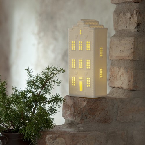 Biała ceramiczna dekoracja w kształcie domku z ciepłym światłem w oknach, ustawiona na ceglanej półce obok zielonej gałązki; subtelne oświetlenie i spokojna, domowa atmosfera.