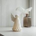 Biała ceramiczna dekoracja LED w kształcie anioła, świecąca delikatnym światłem, ustawiona na jasnej powierzchni obok szklanej butelki z białymi piórami; jasne tło i harmonijna kompozycja.