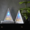 Dwie trójkątne białe dekoracje stołowe LED, jedna z motywem szopki bożonarodzeniowej, druga z zimowym krajobrazem. Obie świecą ciepłym i niebieskim światłem, ustawione na komodzie obok zielonej rośliny, tworzą harmonijną, przytulną kompozycję.