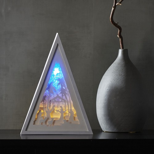 Trójkątna biała dekoracja stołowa LED przedstawiająca szopkę bożonarodzeniową z warstwową sceną Narodzenia, świecąca ciepłym i niebieskim światłem, ustawiona na blacie obok wazonu. Stonowane tło i spokojna atmosfera.