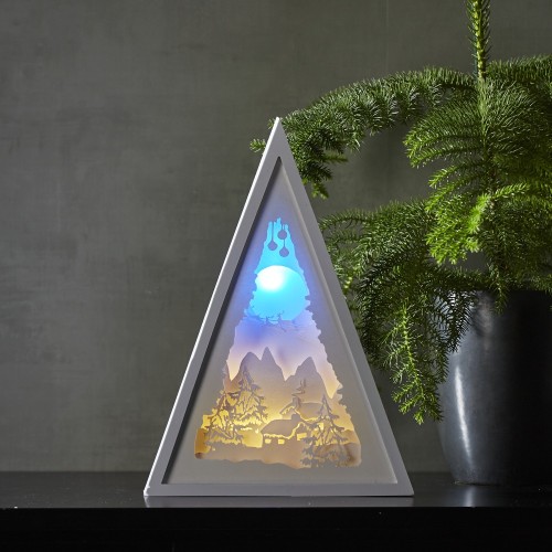 Trójkątna biała dekoracja stołowa LED z motywem zimowego krajobrazu gór, drzew i domu w śnieżnej scenerii, świecąca ciepłym i niebieskim światłem, ustawiona obok zielonej rośliny. Spokojna atmosfera.