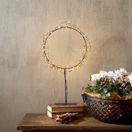 Dekoracyjny wieniec LED osadzony na smukłej podstawie, ustawiony na drewnianym meblu; ciepłe punkty światła, w tle książki i misa z białymi kwiatami. Spokojna, przytulna ozdoba do wnętrza.