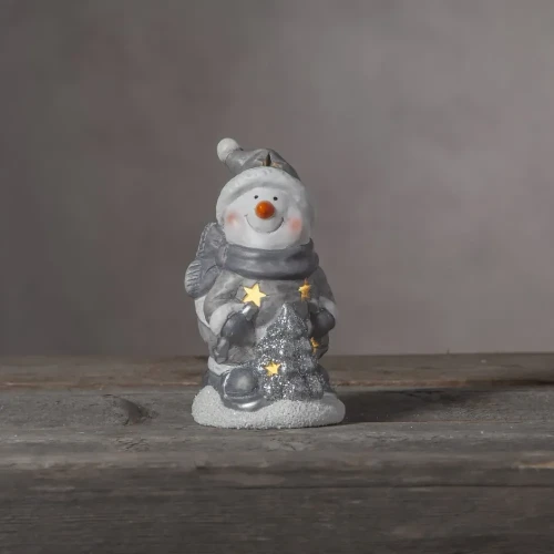 Figurka bałwanka w szaro-białej kolorystyce, trzymającego choinkę ze świecącymi gwiazdkami, ustawiona na drewnianej powierzchni, dekoracja świąteczna w nastrojowym, przytulnym klimacie.