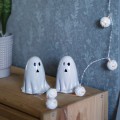 Dwie ceramiczne figurki duszków na drewnianej półce przy ścianie, udekorowane girlandą świetlną z lampkami w kształcie dyni. Ciepłe punkty światła tworzą spokojną, dekoracyjną scenę.