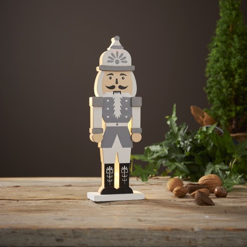 Drewniana figurka dziadka do orzechów z podświetleniem LED, stojąca na rustykalnym stole obok zielonej rośliny i orzechów. Ciepłe światło tworzy spokojny, świąteczny klimat.