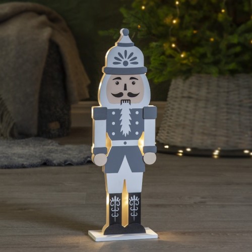 Drewniana figurka dziadka do orzechów z podświetleniem LED, stojąca na podłodze w przytulnym wnętrzu, obok pledu i choinki z lampkami. Ciepłe światło buduje spokojny, świąteczny klimat.