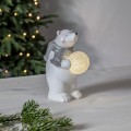 Biały miś z brokatowym wykończeniem i srebrnym szalikiem, trzymający świecącą śnieżną kulę LED, stojący na jasnym blacie. W tle choinka z lampkami tworzy spokojny, świąteczny klimat.