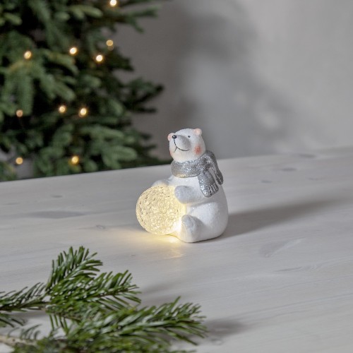 Biały miś z brokatowym wykończeniem i srebrnym szalikiem, siedzący na jasnym blacie i trzymający świecącą śnieżną kulę LED. W tle choinka z lampkami buduje spokojny, świąteczny klimat.