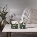 Dwie zielone ceramiczne domki z białymi dachami, ustawione na jasnym stoliku w salonowej aranżacji. W tle miękkie tekstylia i roślina w wazonie, tworzące spokojny, domowy klimat.