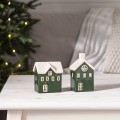 Dwa zielone ceramiczne domki z białymi dachami na jasnym stoliku. W tle rozmyta choinka z lampkami i jasne tekstylia, budujące przytulną, świąteczną aranżację wnętrza.