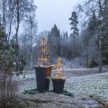 Dwie choinki świetlne LED na metalowym stelażu w czarnych donicach ustawione na zewnątrz w zimowym otoczeniu. Lampki świecą ciepłym światłem, w tle widoczne drzewa oraz lekko ośnieżona trawa.