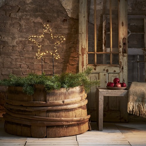 Gwiazda LED na metalowym stelażu z ciepłym światłem, ustawiona w drewnianej beczce z zielonymi gałązkami. Przytulna dekoracja świąteczna we wnętrzu w rustykalnym stylu, na tle ceglanej ściany.