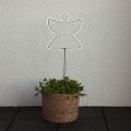 Anioł NEON LED na metalowym pręcie, świecący jasnym światłem, umieszczony w doniczce z zieloną rośliną. Dekoracyjna ozdoba świetlna do wnętrz, zaprezentowana na tle jasnej ściany.
