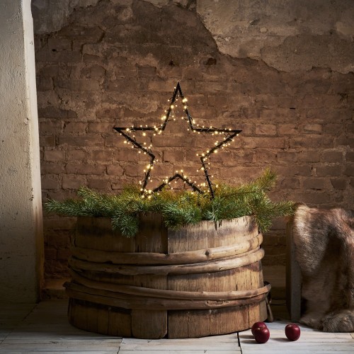 Dekoracyjna gwiazda LED z drobnymi punktami świetlnymi ustawiona w drewnianej beczce z zielonymi gałązkami; ciepłe światło na tle surowej, ceglanej ściany.