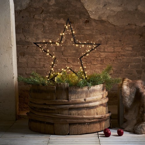 Duża dekoracyjna gwiazda LED z drobnymi punktami świetlnymi ustawiona w drewnianej beczce z gałązkami iglastymi; ciepłe światło tworzące przytulną atmosferę na tle ceglanej ściany.
