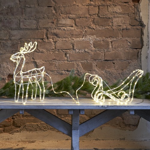 Zestaw dekoracji świetlnych LED z saniami i reniferami ustawiony na drewnianej ławie; ciepłe światło tworzy przytulną świąteczną aranżację na tle ceglanej ściany.