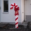 Podświetlana dekoracja świąteczna LED w kształcie czerwono-białej laski cukrowej z dużą czerwoną kokardą, ustawiona przed domem na metalowej podstawie, emitująca ciepłe, nastrojowe światło.