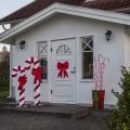 Dekoracja LED w kształcie czerwonego prezentu z czerwono-białymi laskami cukrowymi zdobi wejście do domu obok świetlnych lasek cukrowych oraz czerwonej kokardy zawieszonej na drzwiach.