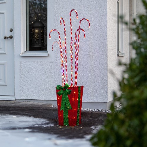 Świąteczna dekoracja LED z czerwono-białymi laskami cukrowymi umieszczonymi w ozdobnej podstawie w formie prezentu z zieloną kokardą, ustawiona przed wejściem do domu.