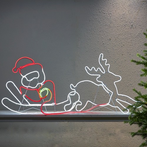 Dekoracja świetlna neon LED przedstawiająca Świętego Mikołaja na saniach z reniferem, ustawiona na półce przy ścianie. Białe, czerwone i zielone światło tworzy wyraźny kontur sceny o świątecznym charakterze.