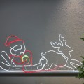 Dekoracja neon LED przedstawiająca Mikołaja na saniach z reniferem, zamontowana przy ścianie obok choinki. Stonowane, jasne światło tworzy wyraźny kontur i przytulny, świąteczny nastrój.
