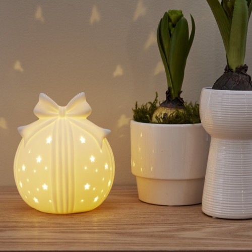 Świecąca ceramiczna bombka-prezent z kokardą, ustawiona na blacie obok białych donic z roślinami; ciepłe światło LED subtelnie rozświetla otwory w kształcie gwiazdek i odbija się na ścianie.