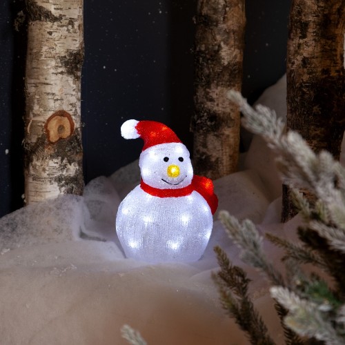 Podświetlana figurka LED bałwanka w czerwonej czapce i szaliku, ustawiona na śniegu między pniami drzew, zimowa aranżacja dekoracyjna z białym światłem.