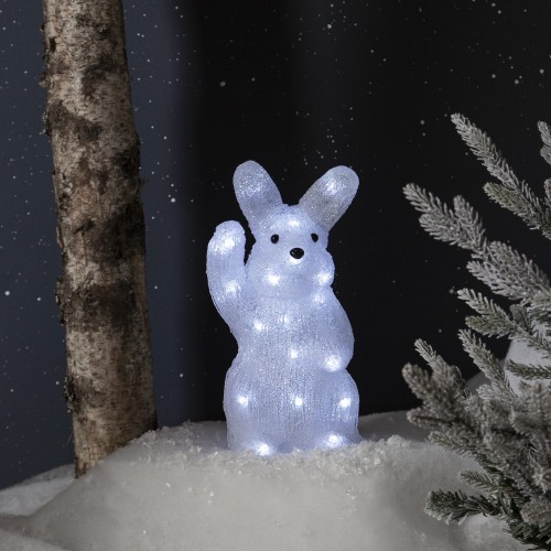 Podświetlana figurka LED w kształcie białego zająca stojącego na śniegu. Dekoracja umieszczona w zimowej aranżacji z pniem drzewa i ośnieżonymi gałązkami w tle.