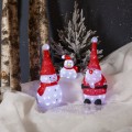 Podświetlane figurki LED ustawione na śniegu: dwa bałwanki oraz Mikołaj w czerwonym stroju. W tle pnie drzew, ośnieżone drzewko i zimowa sceneria.