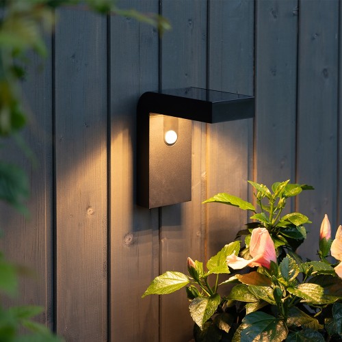 Czarny solarny kinkiet ogrodowy LED VICI z czujnikiem ruchu PIR zamontowany na drewnianej elewacji ciepłe światło oświetlające rośliny przy ścianie.