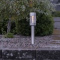 Solarny słupek ogrodowy LED wbijany w ziemię, lampa solarna zewnętrzna ustawiona na żwirze przy rabacie i krzewach, delikatne światło dekoracyjne do ogrodu.