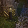 Czarna lampa solarna LED przy drzewie i kamiennej nawierzchni. Perforowana obudowa tworzy dekoracyjne punkty światła na trawie i pniu. Oświetlenie ogrodowe do podkreślenia ścieżek i przestrzeni wokół domu.