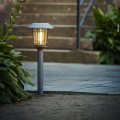 Lampa solarna ogrodowa w formie słupka z daszkiem i przeszkloną osłoną, emitująca ciepłe światło. Ustawiona w ziemi przy kamiennych schodach i roślinach, delikatnie rozświetla przestrzeń po zmroku.