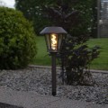 Lampa ogrodowa solarna wbijana z dekoracyjną żarówką LED w stylu filament świeci ciepłym światłem w rabacie z kamieniami i niskimi krzewami o zmierzchu.